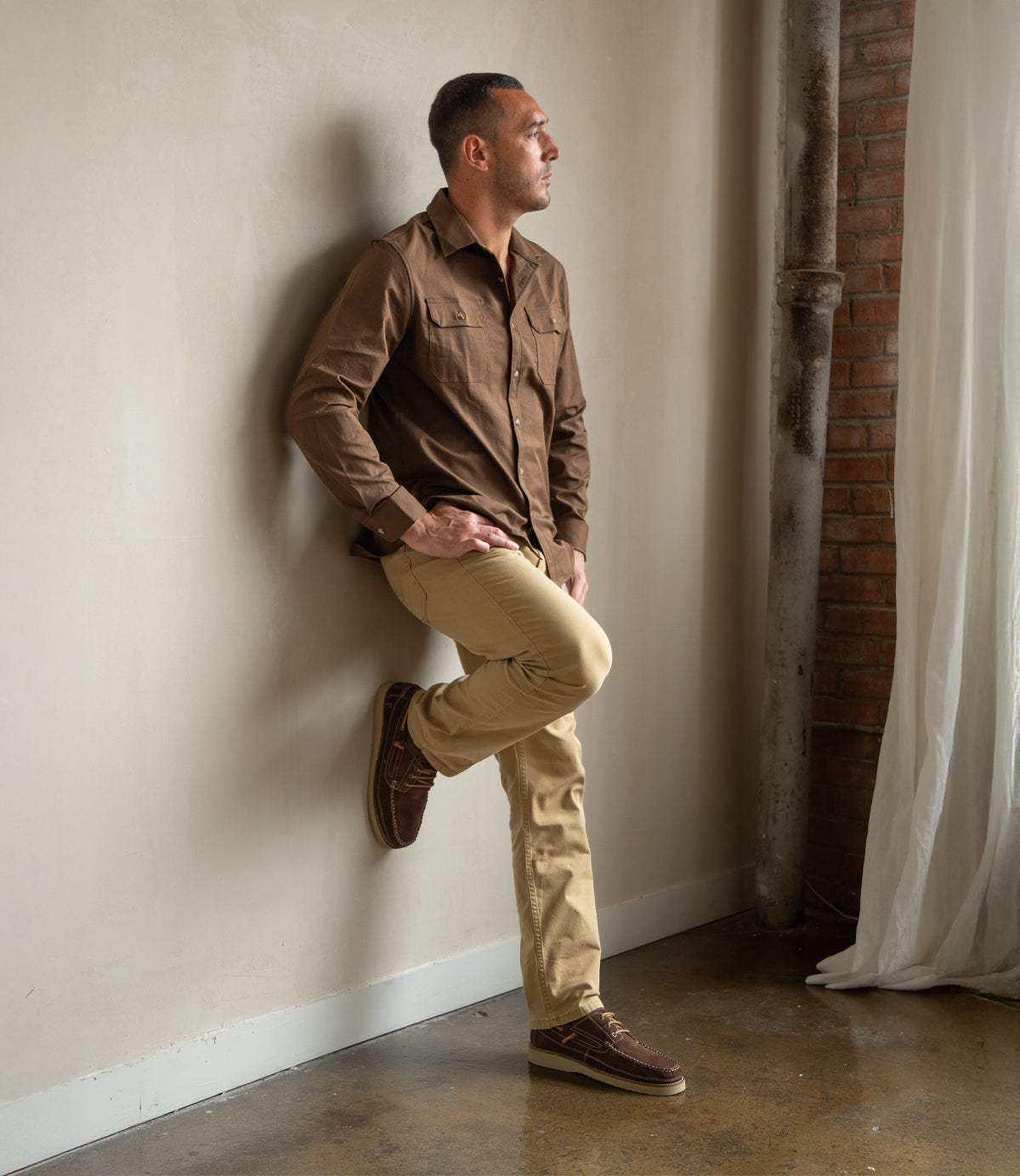 A woman in a white sweater, blue jeans, mustard beanie, and yellow socks sits sideways on a wooden chair by a brick wall and large windows, wearing the Roan Abide durable loafer.