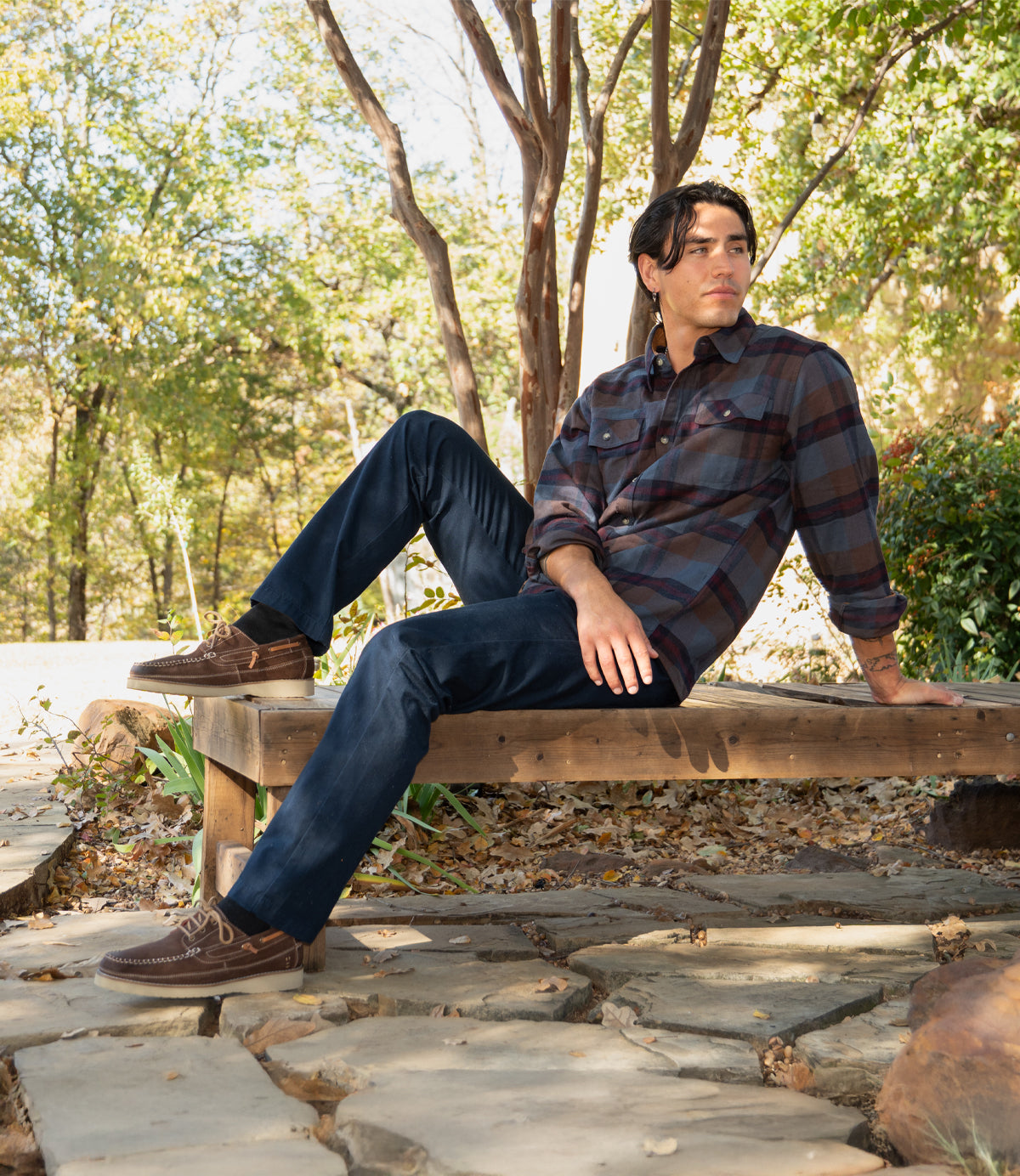 A man in a plaid shirt and dark jeans, wearing Roan's Glory lace-up boat shoes, sits on a wooden bench outdoors among trees and stone pavement.