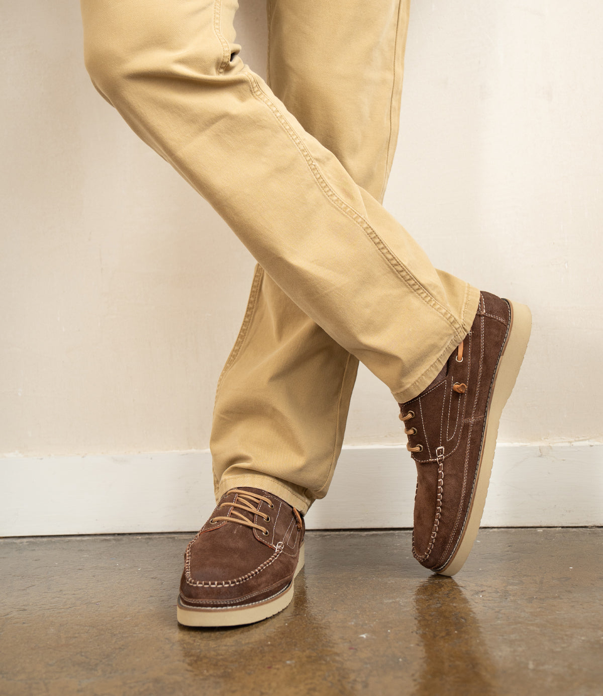 A person wearing Roan's Glory tan pants and brown suede boat shoes stands with legs crossed on a polished concrete floor.