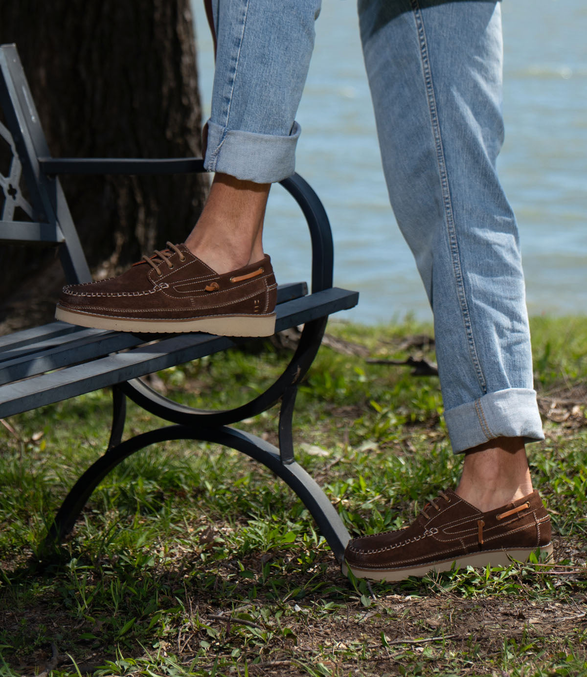 A person wearing Roan's Glory brown suede boat shoes with cushioned footbed and cuffed light blue jeans stands on a park bench by grass and water.