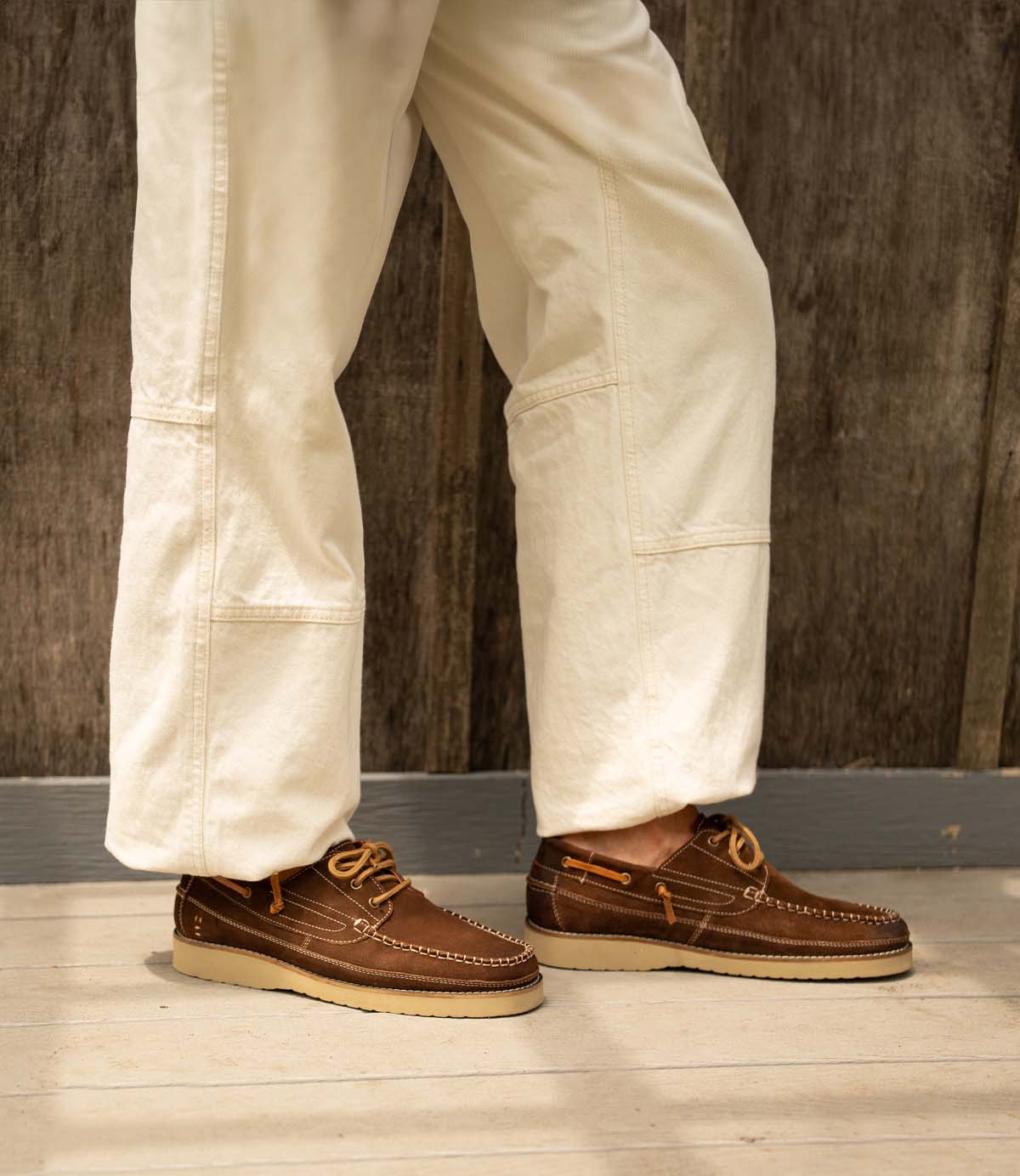 A person in white pants wears Roan's Glory brown leather boat shoes with a cushioned footbed, standing on a wooden floor before a rustic wooden wall.