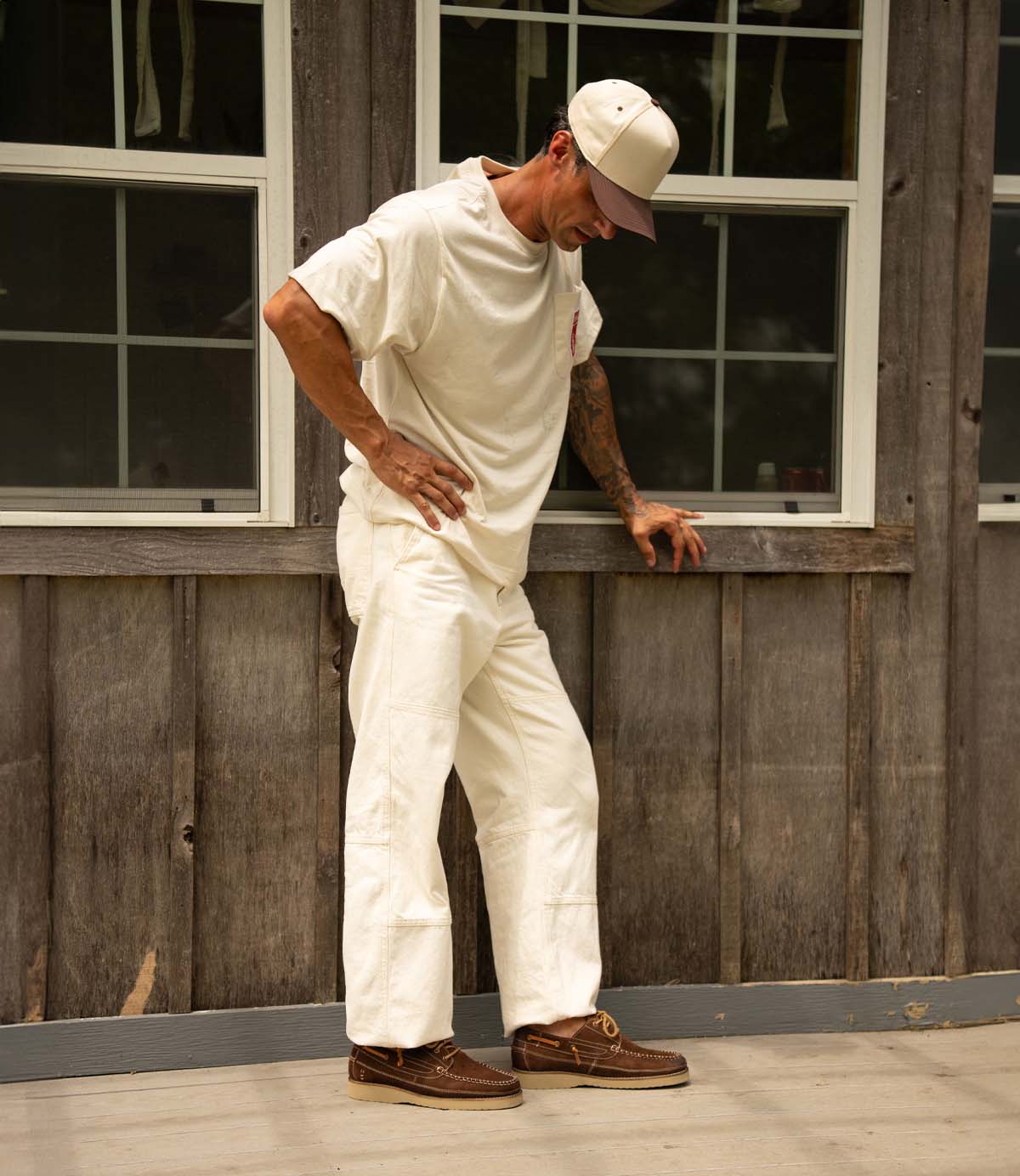 
                  
                    A man stands on a porch wearing the Roan Glory beige cap, an off-white t-shirt, white pants, and classic boat shoes, looking down with one hand on his hip and the other resting on a wooden wall.
                  
                