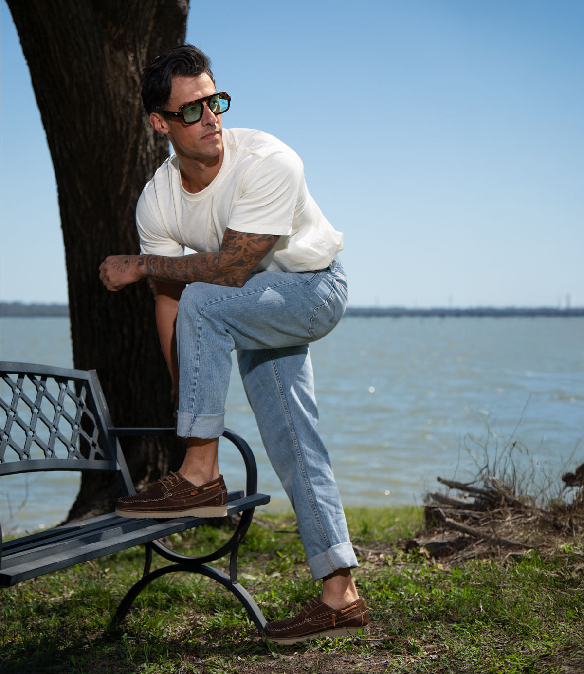
                  
                    A man in a white t-shirt, rolled-up jeans, and sunglasses poses by a lake on a sunny day, showing off his Roan Glory lace-up boat shoes with one foot on a bench.
                  
                