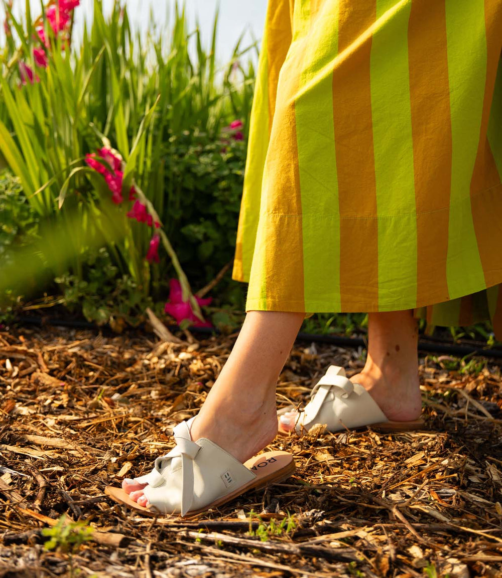 A person walks on a mulch path lined with green plants and pink flowers, wearing Roan's Grapevine yellow and green striped skirt and white leather sandals with cushioned insoles.