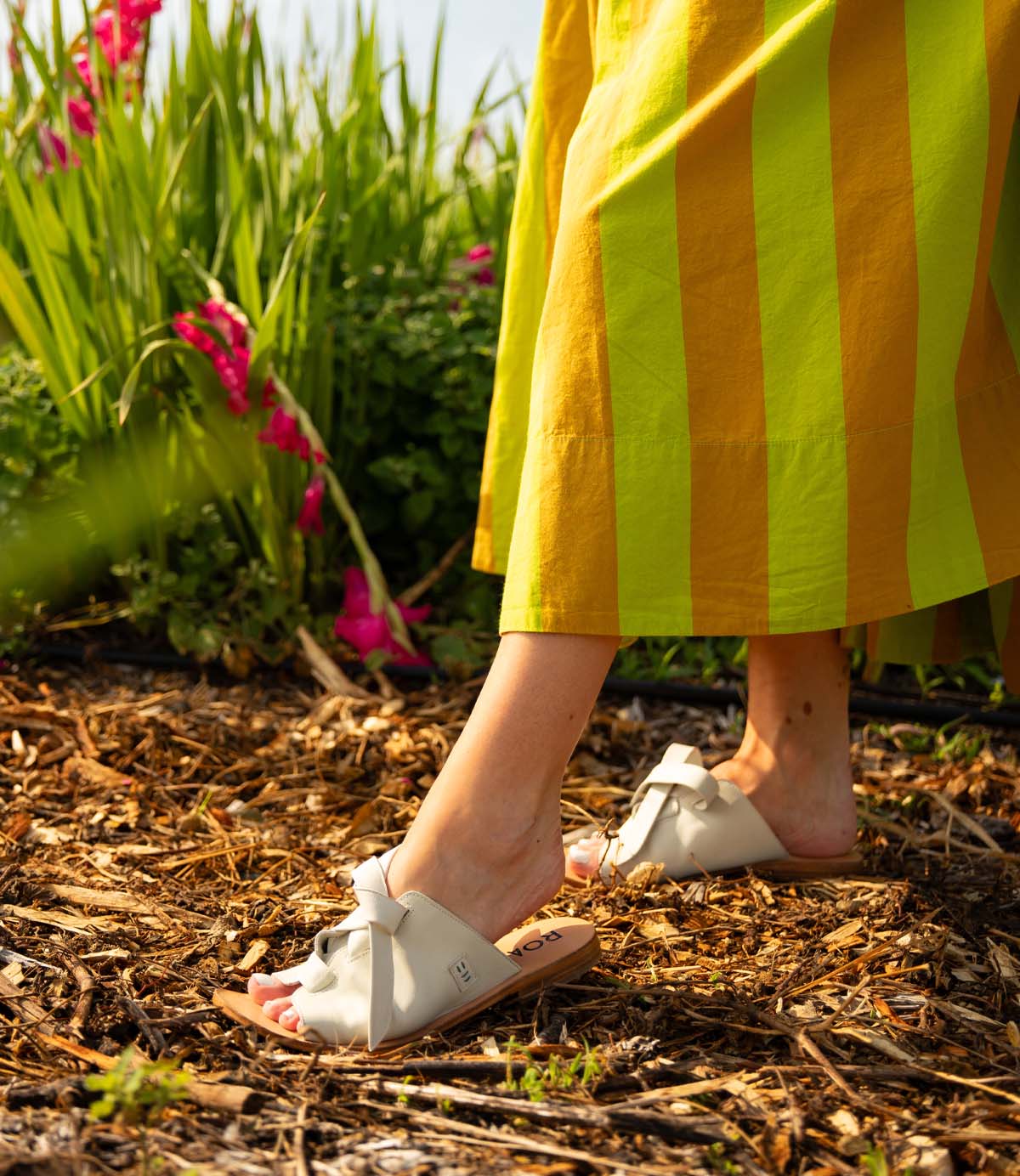 
                  
                    A person walks on a mulch path lined with green plants and pink flowers, wearing Roan's Grapevine yellow and green striped skirt and white leather sandals with cushioned insoles.
                  
                