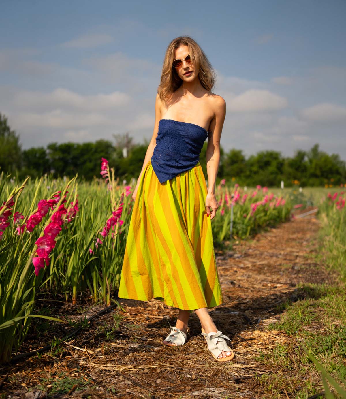 
                  
                    A woman in sunglasses, a blue strapless top, and a yellow striped skirt stands among pink flowers beneath a partly cloudy sky, pairing her look with Roan's Grapevine sandals—stylish leather footwear with adjustable straps.
                  
                