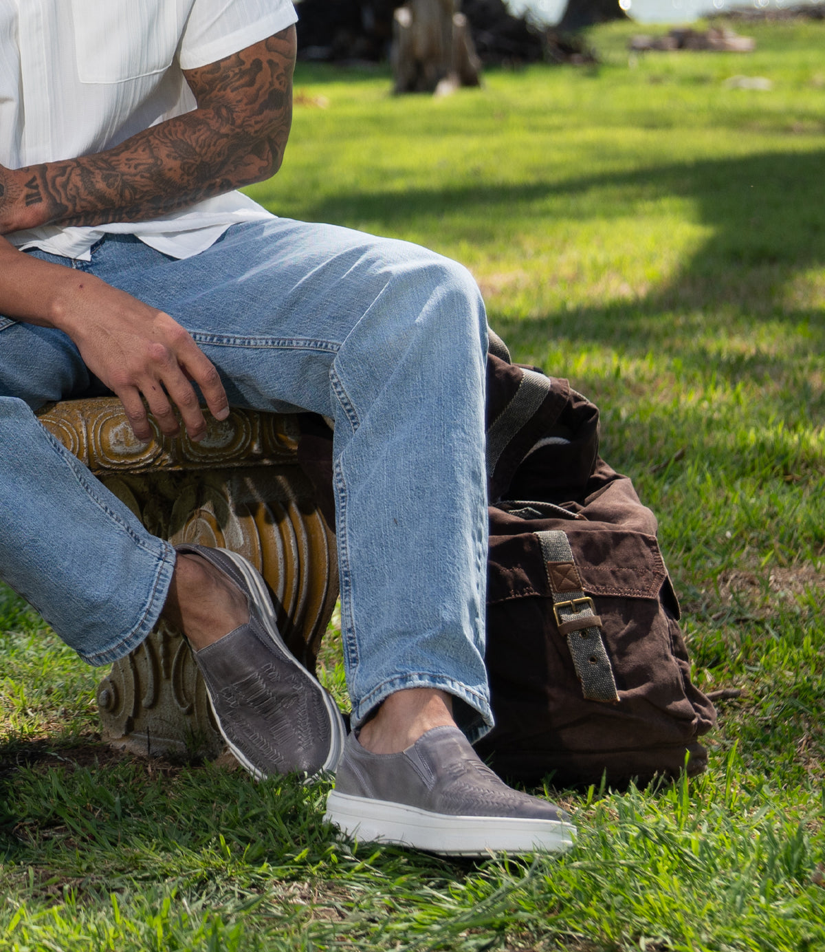 A person wearing Roan Gregor light blue washed jeans and gray slip-on shoes sits on a bench in a grassy area, with a brown backpack on the ground nearby.