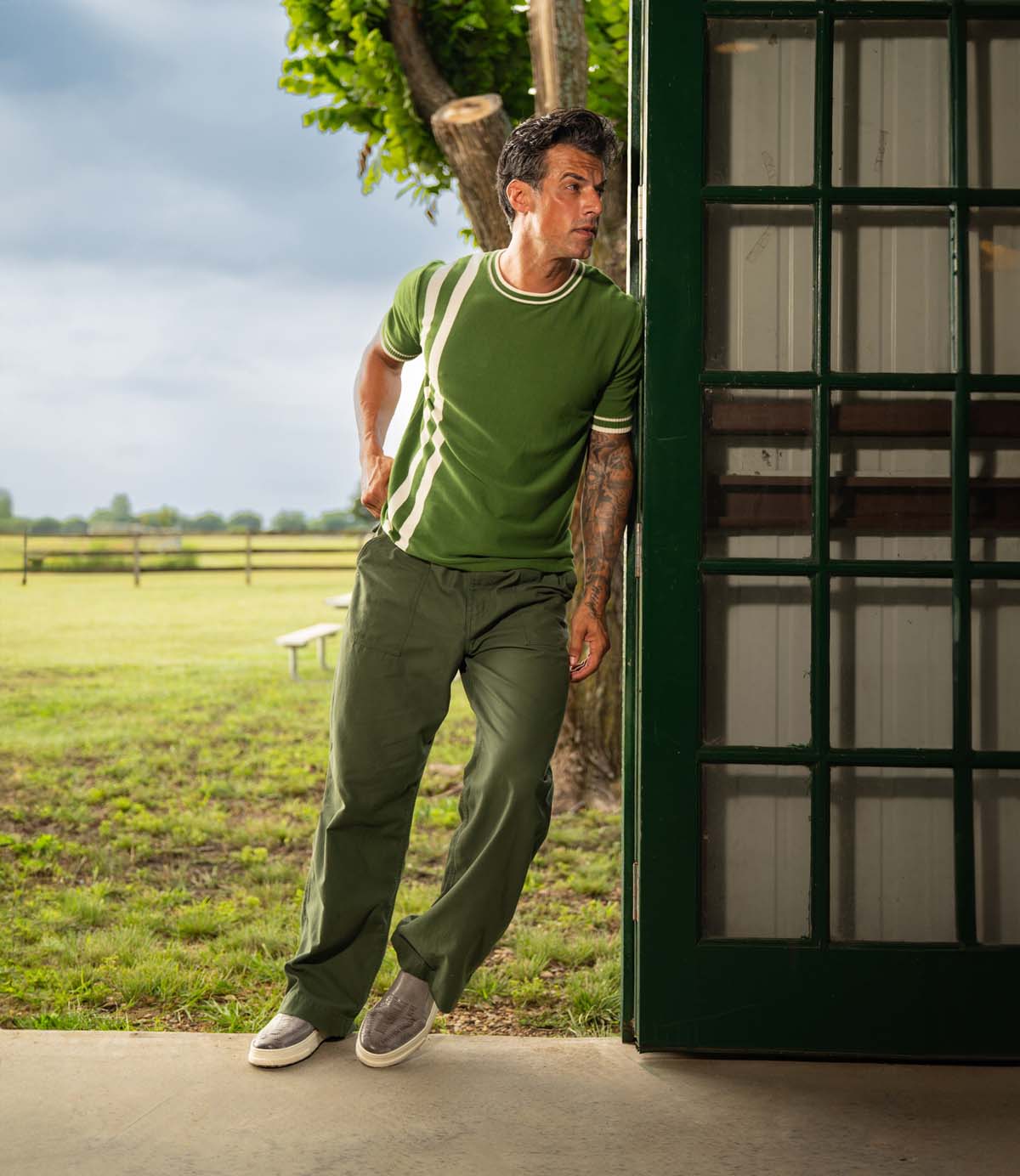 
                  
                    A man wearing the Roan Gregor green shirt and pants with slip-on shoes leans against a green-framed glass door, overlooking a grassy field and trees.
                  
                