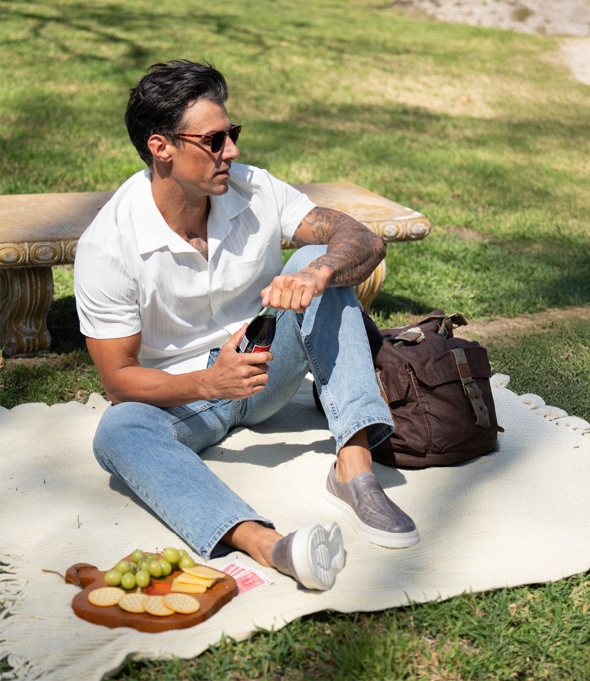 A man in sunglasses and a white shirt relaxes on a blanket outdoors, opening a drink next to cheese, crackers, and grapes on a wooden tray, while wearing stylish Gregor slip-ons by Roan.