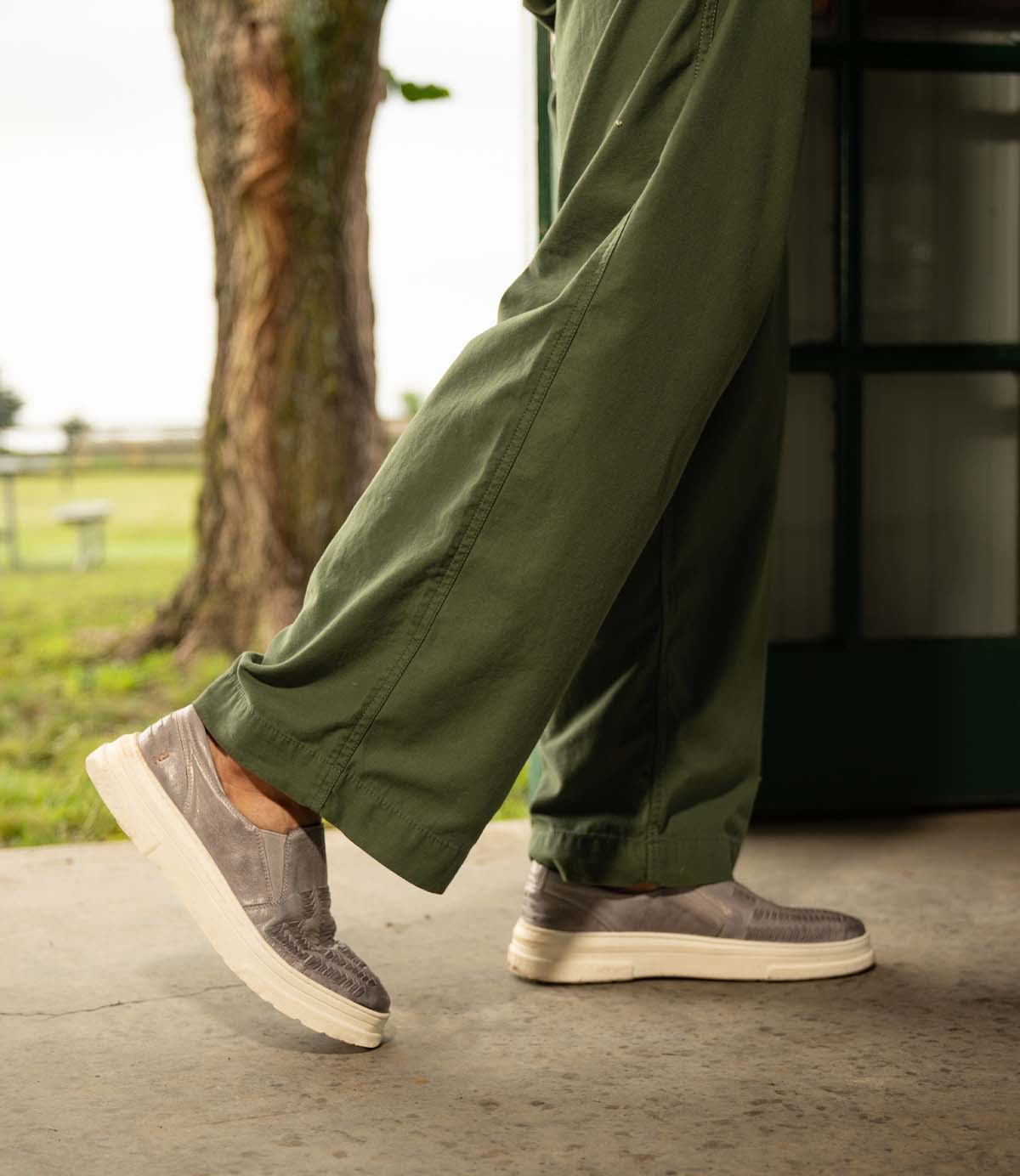 Wearing Roan's Gregor olive green pants and gray washed slip-on sneakers, a person walks on a concrete path outdoors with grass and trees in the background.