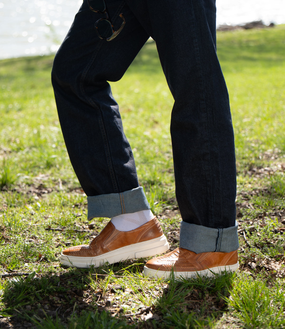 A person wearing Roan's Nosy tan slide sandals with a leather footbed and blue jeans is relaxing, feet up against a peach-patterned geometric wall. This easygoing look captures the essence of summer style perfectly.