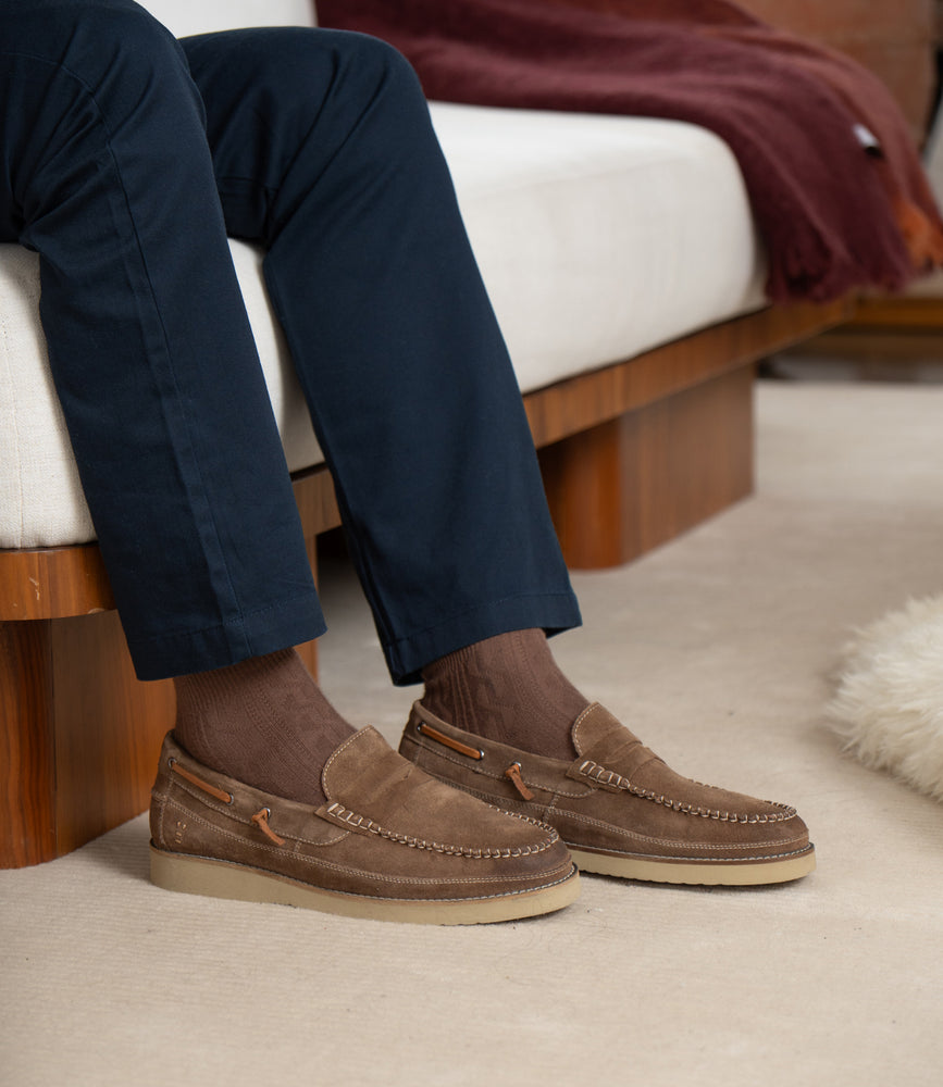 A person wearing Roan Grid brown suede penny loafers with wave-textured rubber soles, brown socks, and navy blue pants sits on a white sofa with a wooden base.