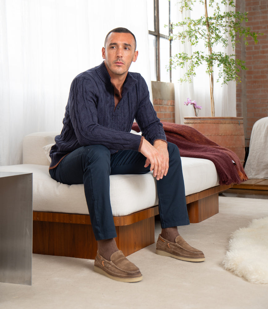 
                  
                    A man sits on Roan’s modern white Grid sofa in a bright living room, wearing a dark cable-knit sweater, navy pants, and suede penny loafers with a wave-textured rubber sole.
                  
                