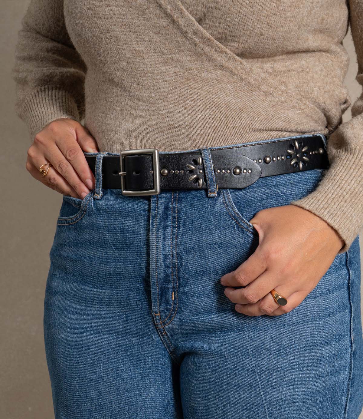 A person wearing a beige sweater, blue jeans, and the Roan “Hollis” black leather strap belt with floral designs and a rectangular buckle, holding the belt and waistband with both hands.