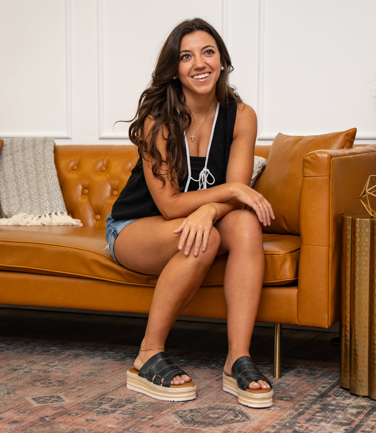 
                  
                    A woman smiles on a tan leather couch, wearing a boho-chic black tank top, denim shorts, and Roan's Incant black platform slide sandals with cushioned footbed. A textured pillow and throw blanket accent the couch.
                  
                