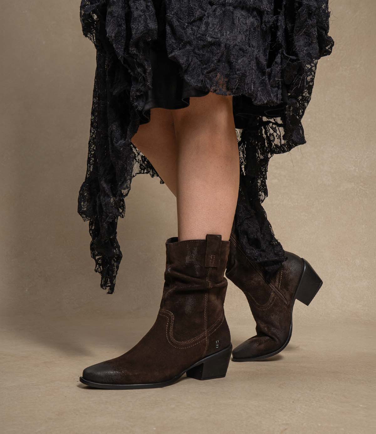 
                  
                    A person is wearing the “Interaction” boots by Roan—dark brown slouchy suede boots featuring a block heel and pointed toe—paired with a black lace skirt, standing against a beige background.
                  
                