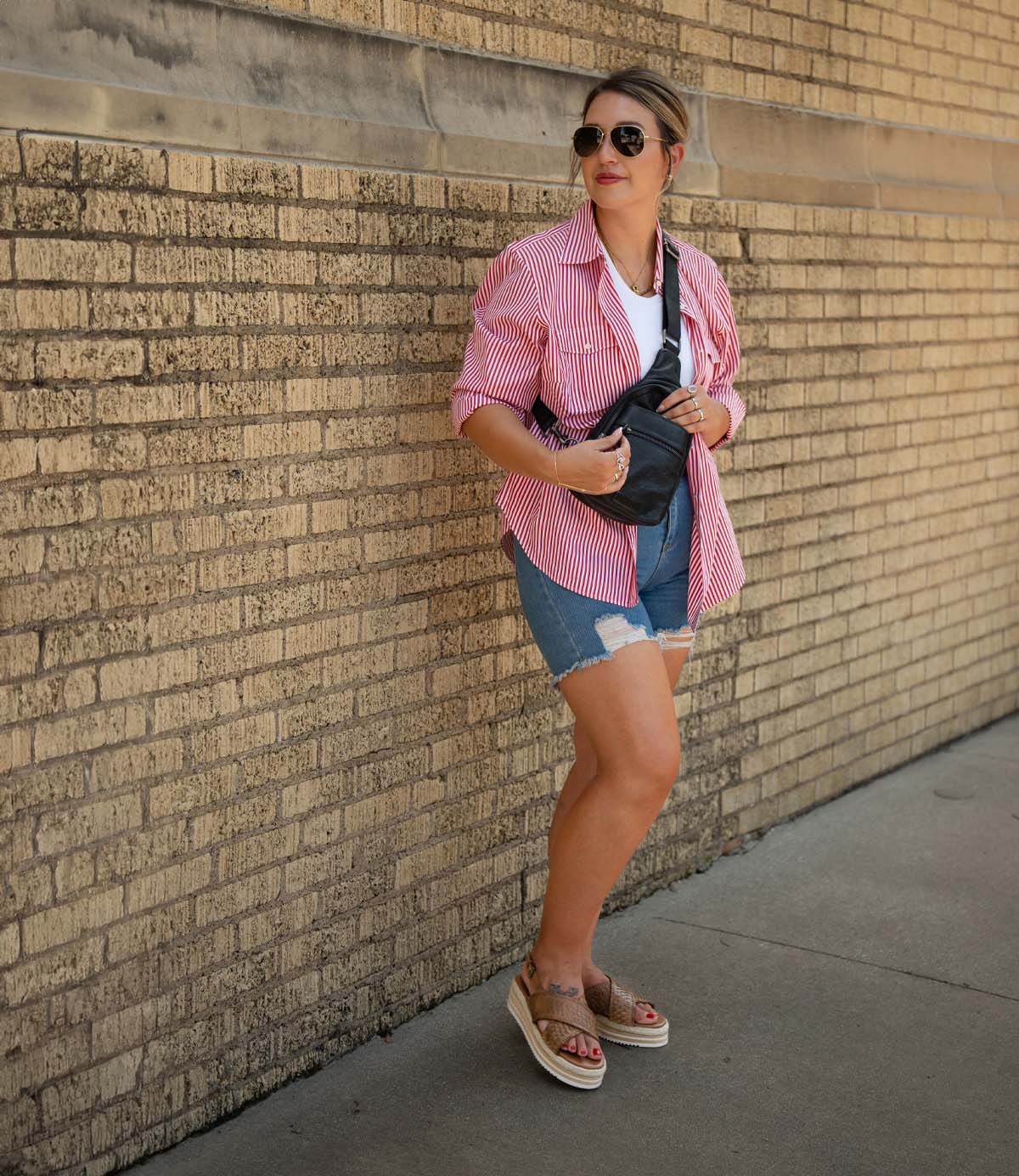 
                  
                    A woman stands against a brick wall wearing the Roan "Into" woven leather sandals, paired with a red striped blazer, white shirt, denim shorts, sunglasses, and a black crossbody bag.
                  
                
