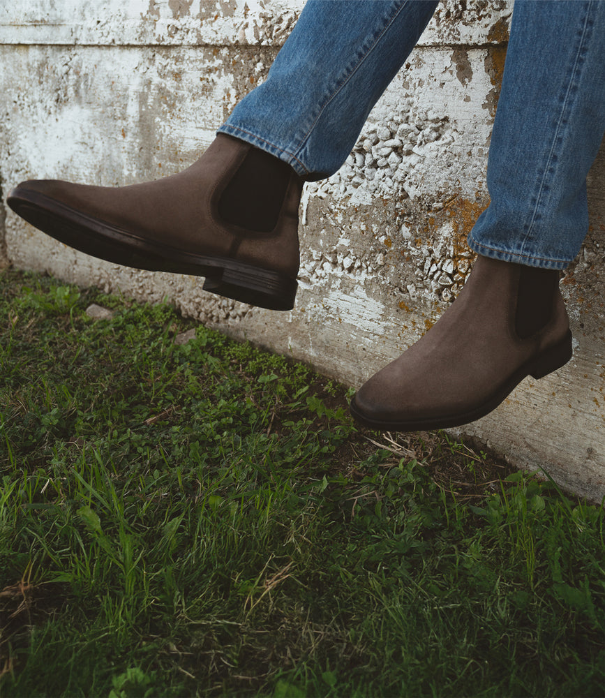 Sporting a pair of Roan's Jardeen, an individual in blue jeans takes pleasure in the slip-on comfort while seated on a concrete ledge overlooking the grass.