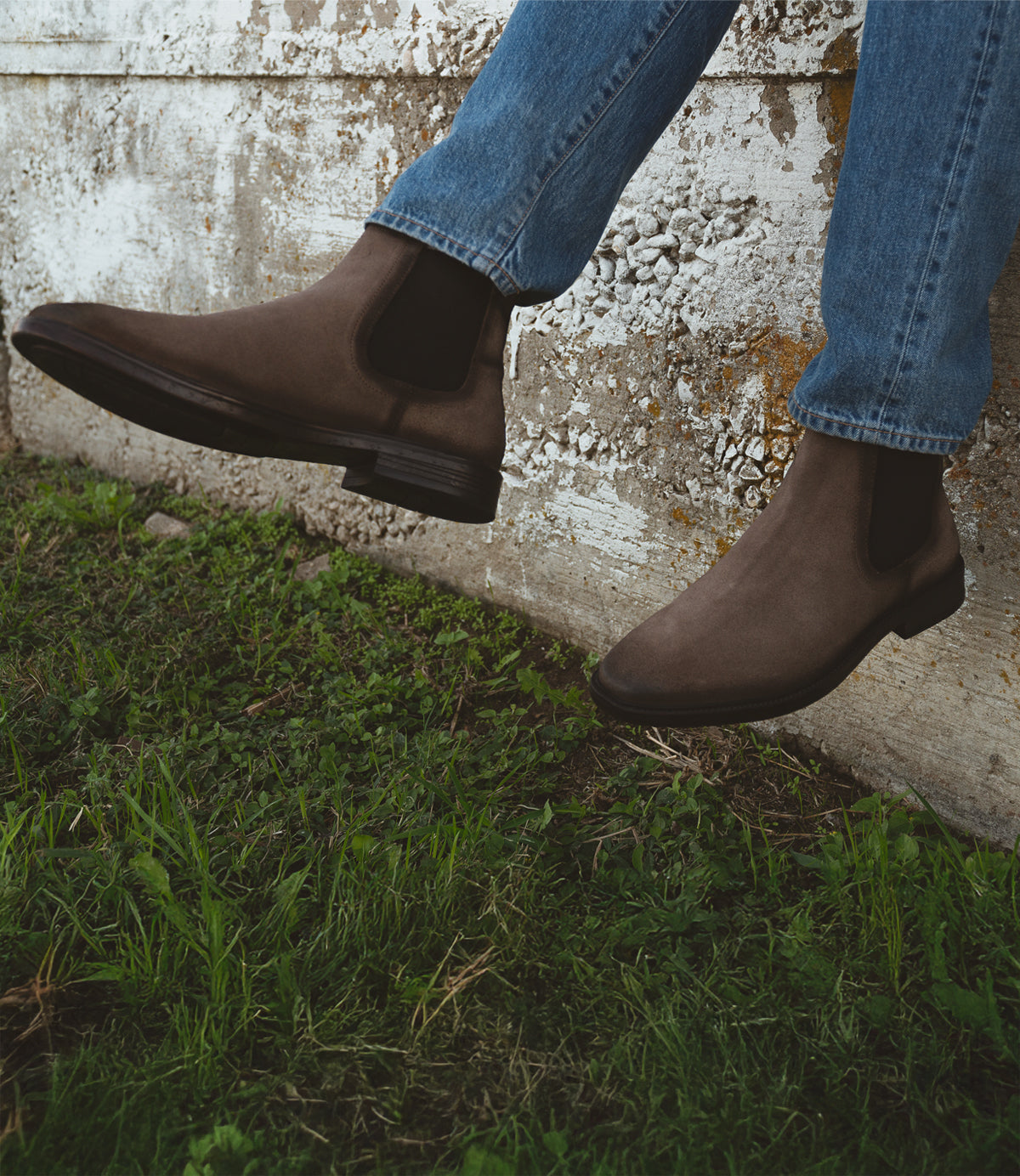Sporting a pair of Roan's Jardeen, an individual in blue jeans takes pleasure in the slip-on comfort while seated on a concrete ledge overlooking the grass.