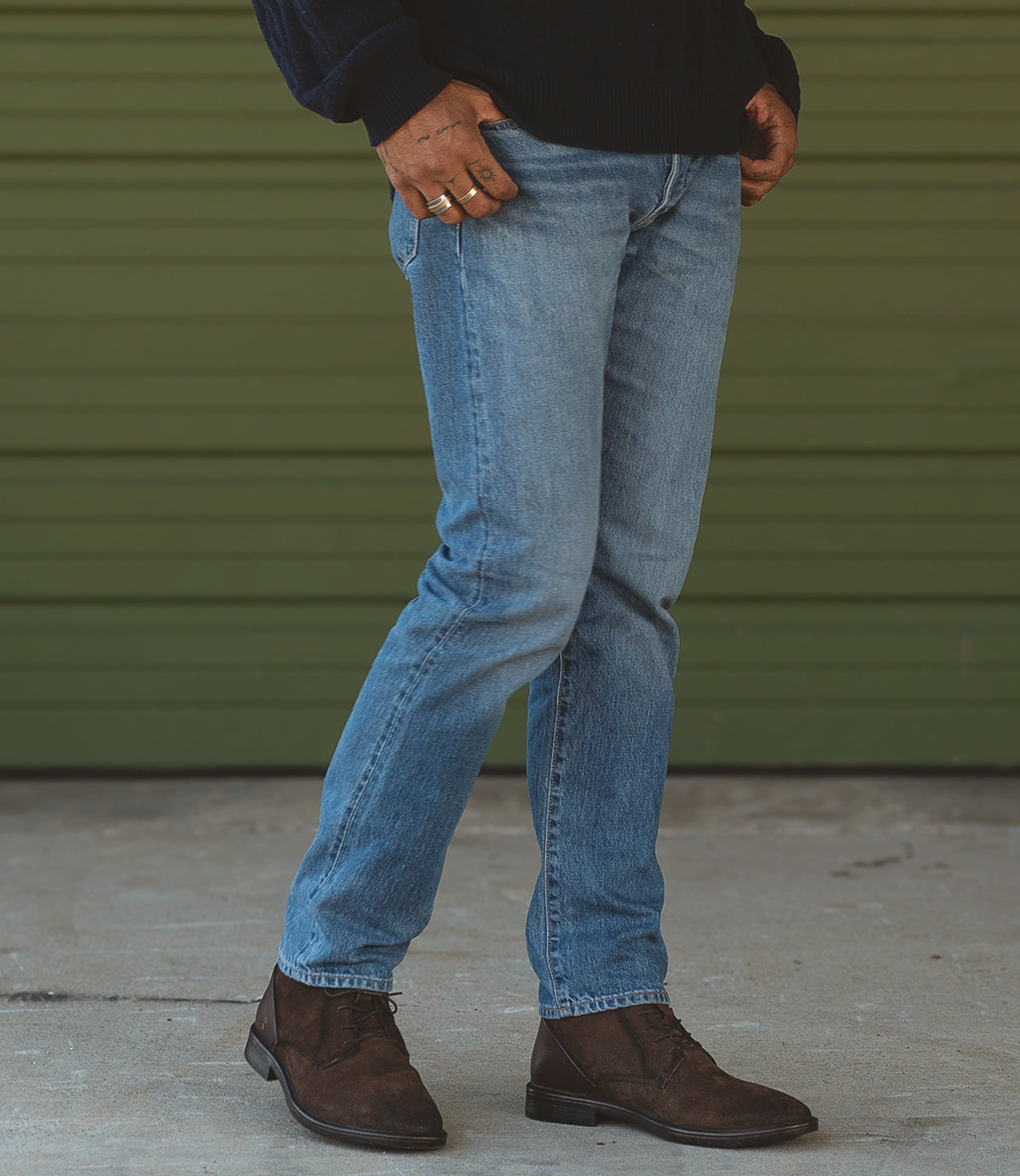 A person wearing Roan's Joyride brown suede lace-up boots and blue jeans stands in front of a green wall.