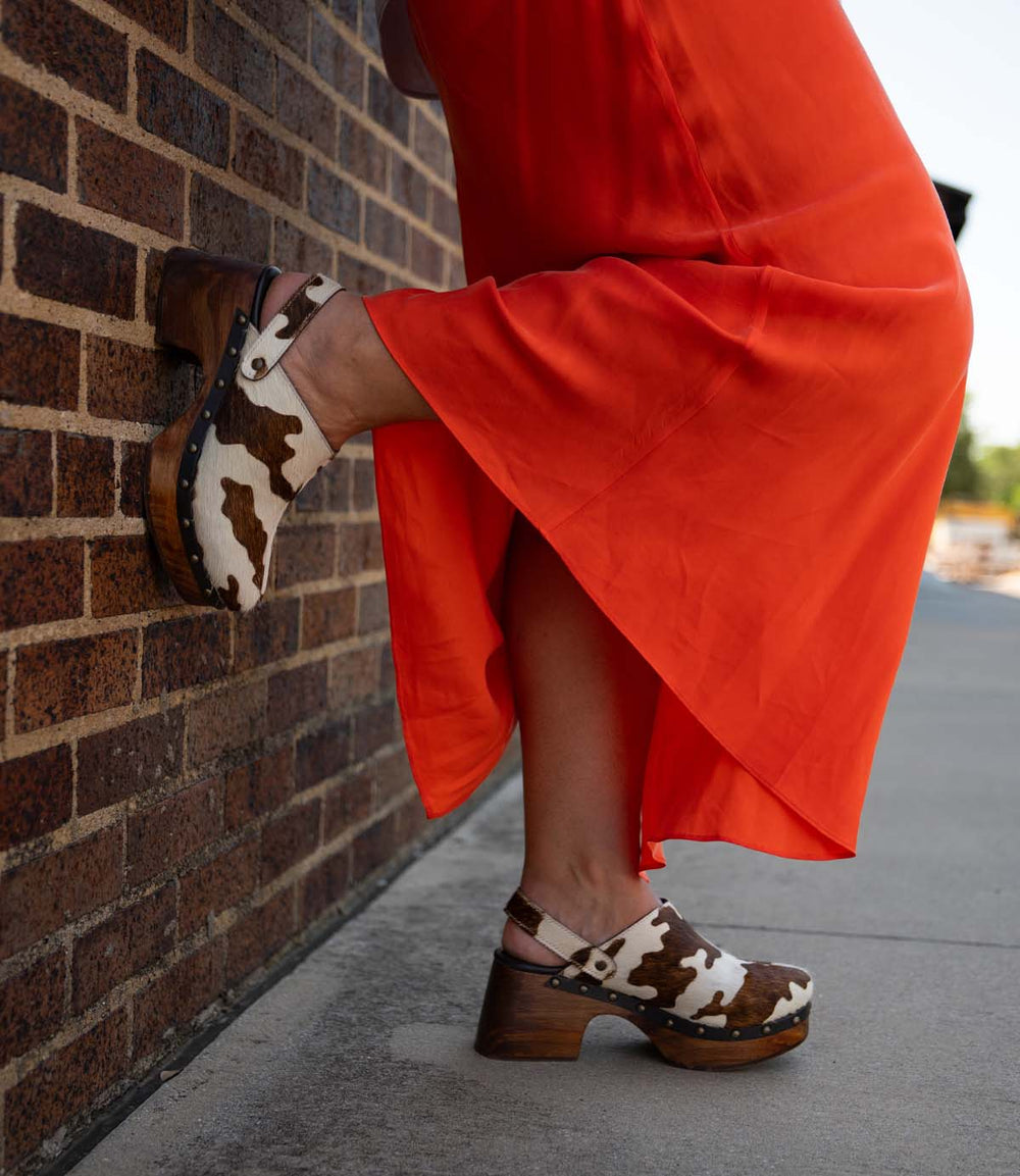 A person in a bright orange dress wears Roan’s Justify brown-and-white hair-on leather clogs with a solid wood base, standing against a brick wall with one foot resting on it.
