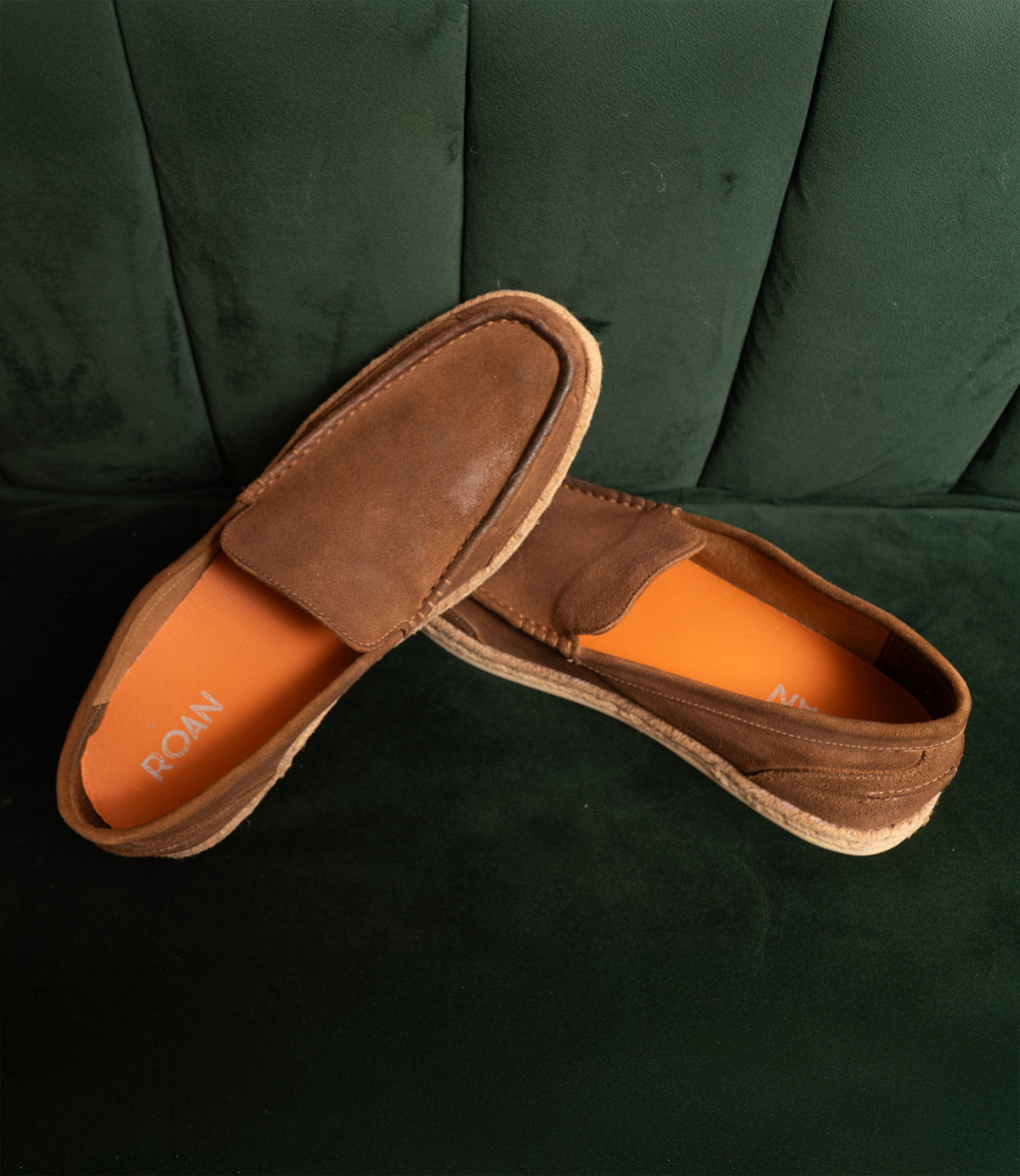 Roan's Woken brown suede loafers, featuring tan soles and orange insoles with hand-finished leather detailing, are elegantly displayed on a dark green cushioned surface.