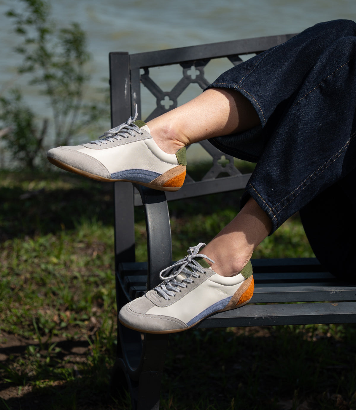 
                  
                    A person sits on a metal bench by grass and water, wearing Roan Keyset low-profile sneakers in white with gray, blue, and orange accents, crafted from premium goat suede for a stylish casual look.
                  
                