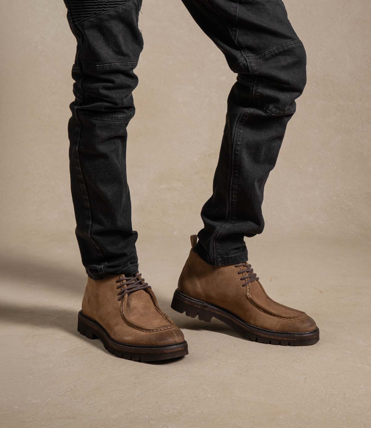 
                  
                    A person wearing black jeans and Roan Kickoff brown suede chukka boots stands on a light-colored floor with a neutral background.
                  
                