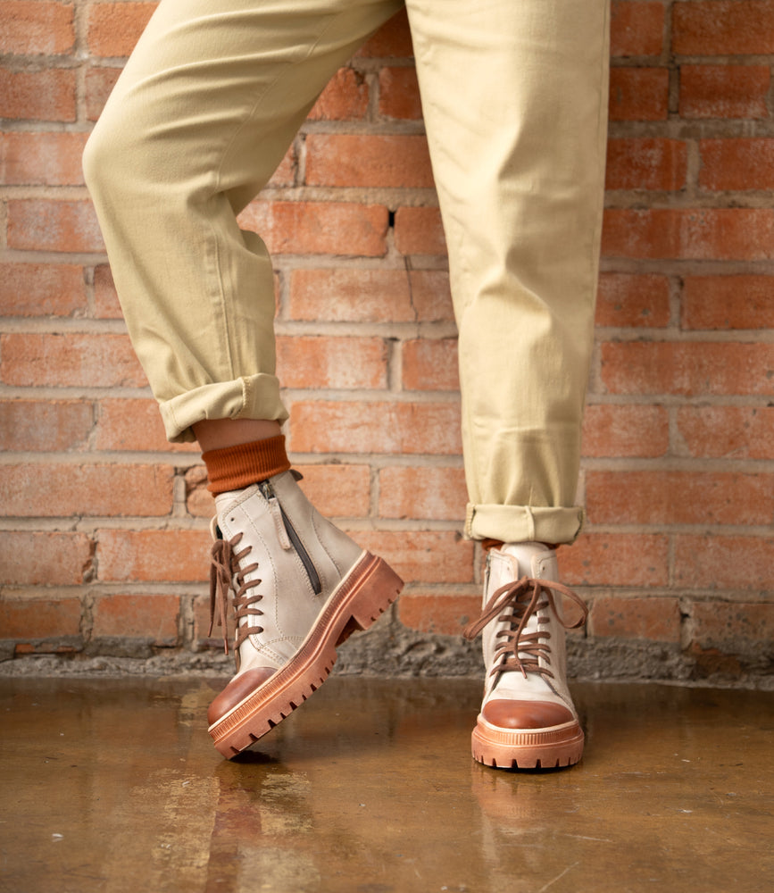 A person wearing beige pants and Roan's Kinship women's leather boots with thick soles stands on a polished floor in front of a brick wall.