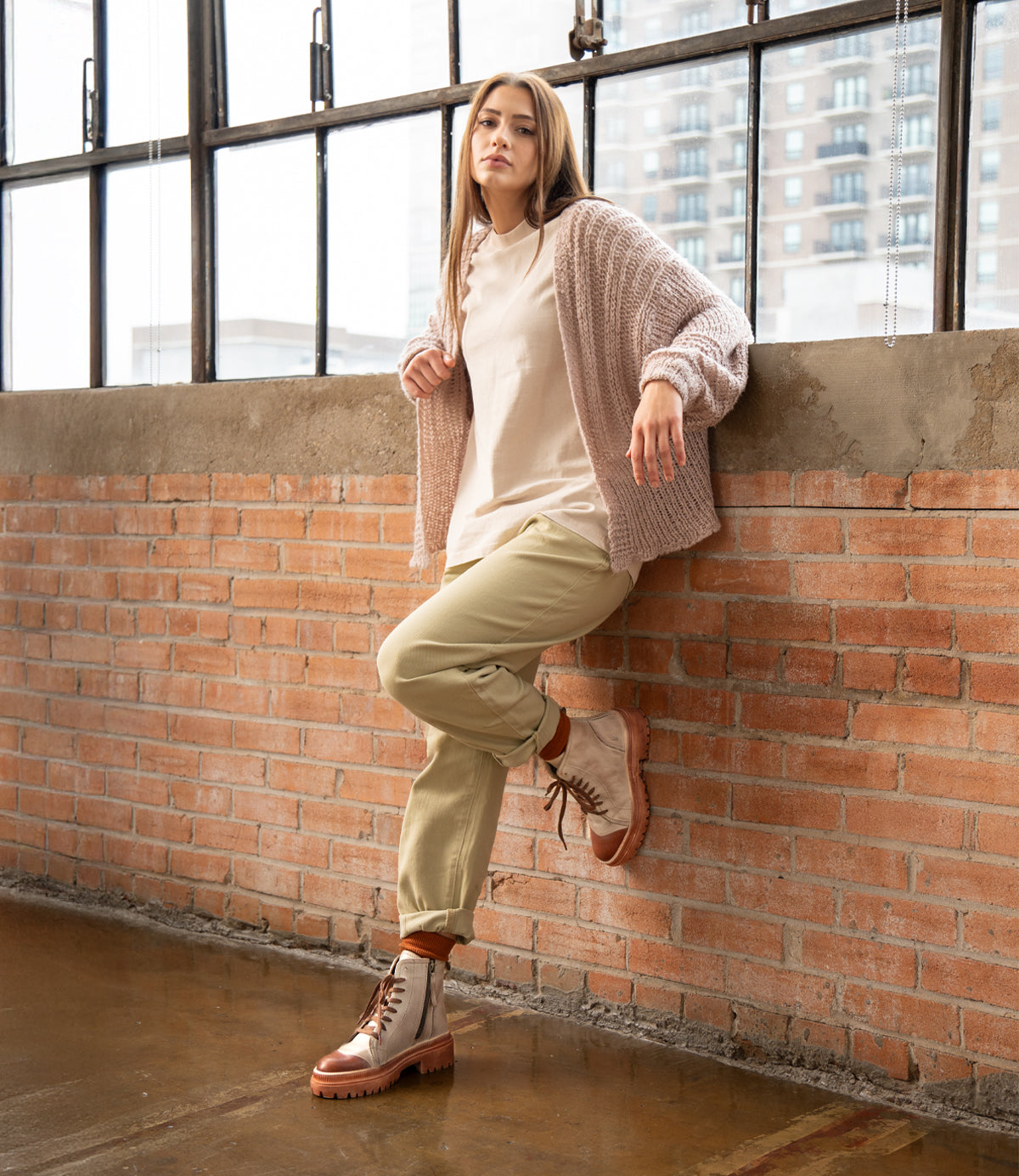 
                  
                    A person wearing Roan's Kinship women's leather boots, a light sweater, and beige pants leans against a brick wall in front of large industrial windows.
                  
                