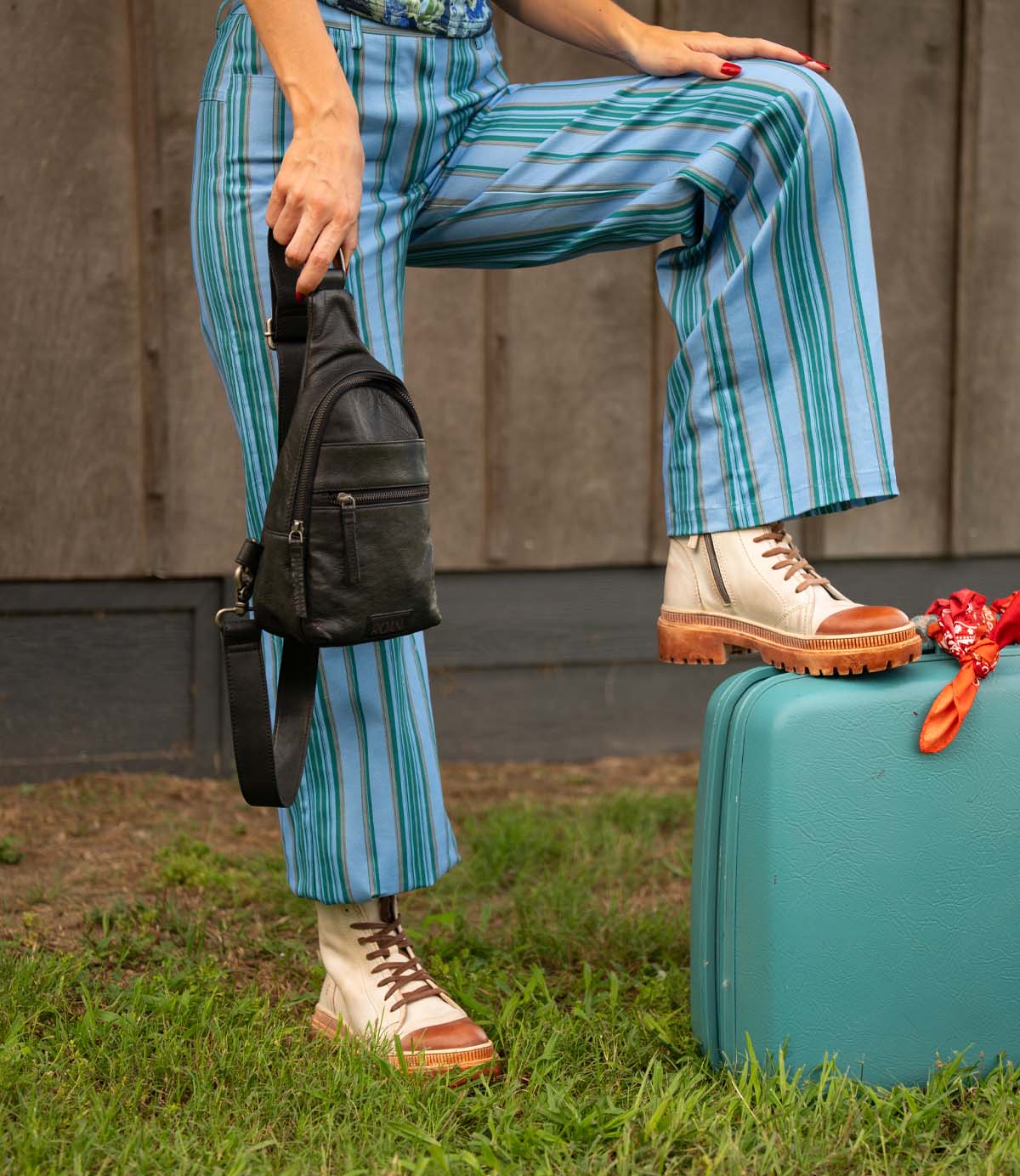 A person stands outdoors with one foot on a teal suitcase, wearing Roan's Kinship blue striped pants and character lace-up leather boots, holding a black leather bag.