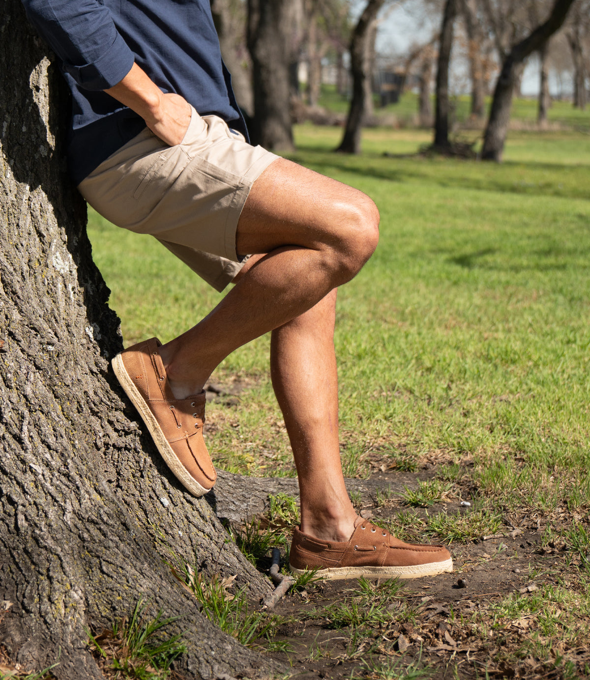 A person wearing Roan Knower tan shorts and casual shoes leans against a tree in a grassy park, one leg bent and hands in pockets.
