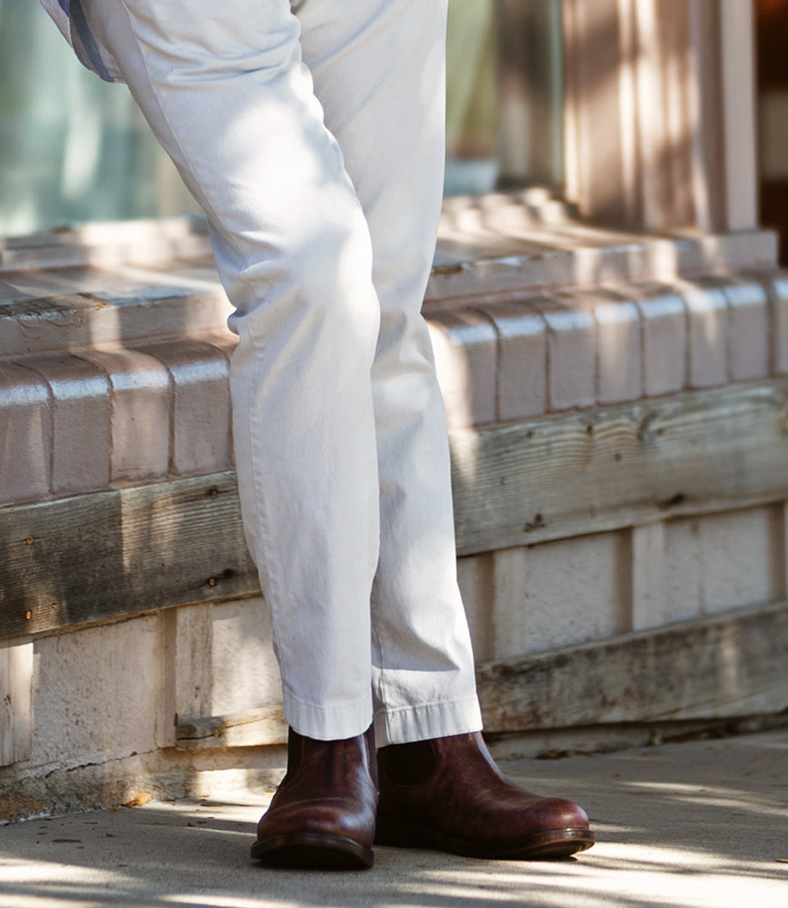 A person wearing the Roan Looting full-grain leather Chelsea boots and white pants stands near a brick ledge, embodying both comfort and style.