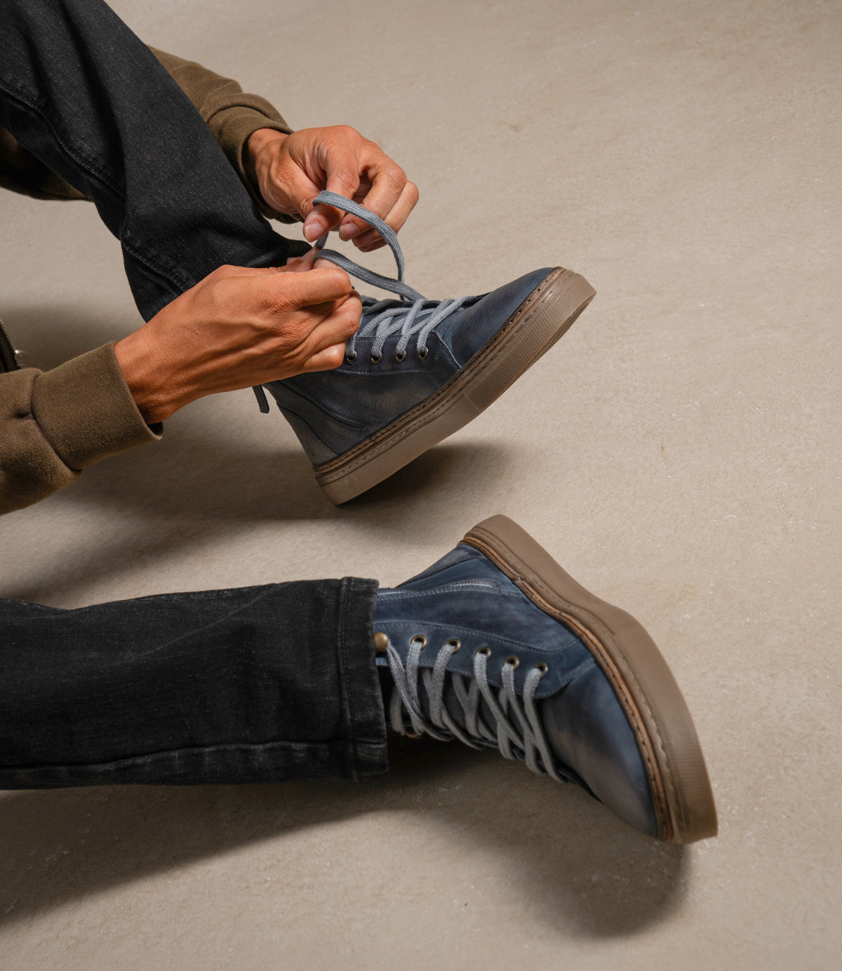 
                  
                    A person sits on the floor tying the laces of blue Roan Mac II high-top leather sneakers with a padded collar, paired with black jeans and a brown sweatshirt.
                  
                