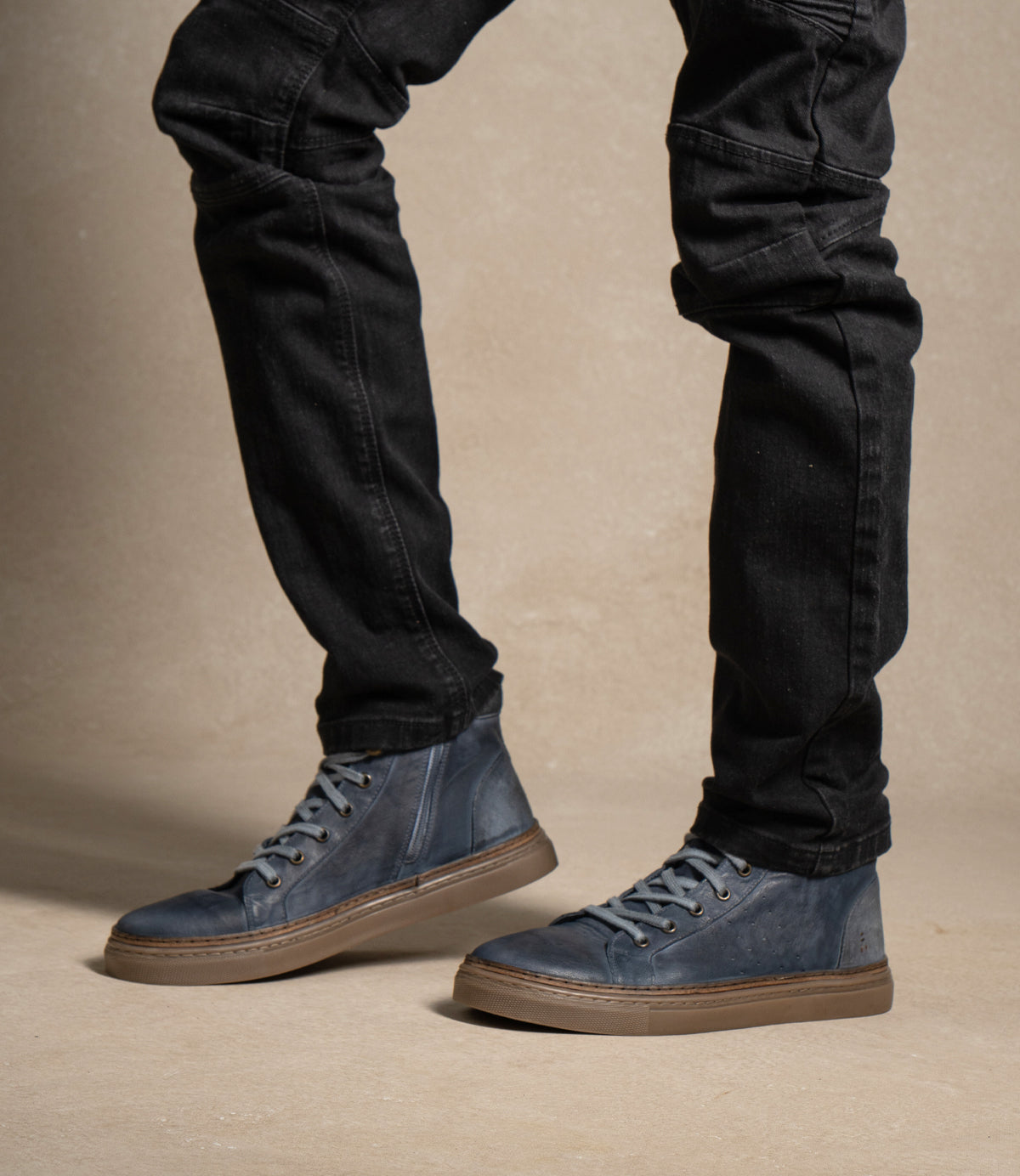
                  
                    A person wearing black jeans and the Roan Mac II blue high-top leather sneakers, featuring a padded collar and brown rubber outsole, stands on a light beige surface against a neutral background.
                  
                