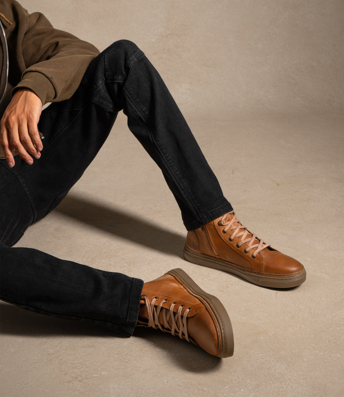 
                  
                    A person sits on the floor wearing black jeans, a brown jacket, and Roan’s Mac II high-top leather sneakers in brown, featuring beige laces and a durable rubber outsole.
                  
                