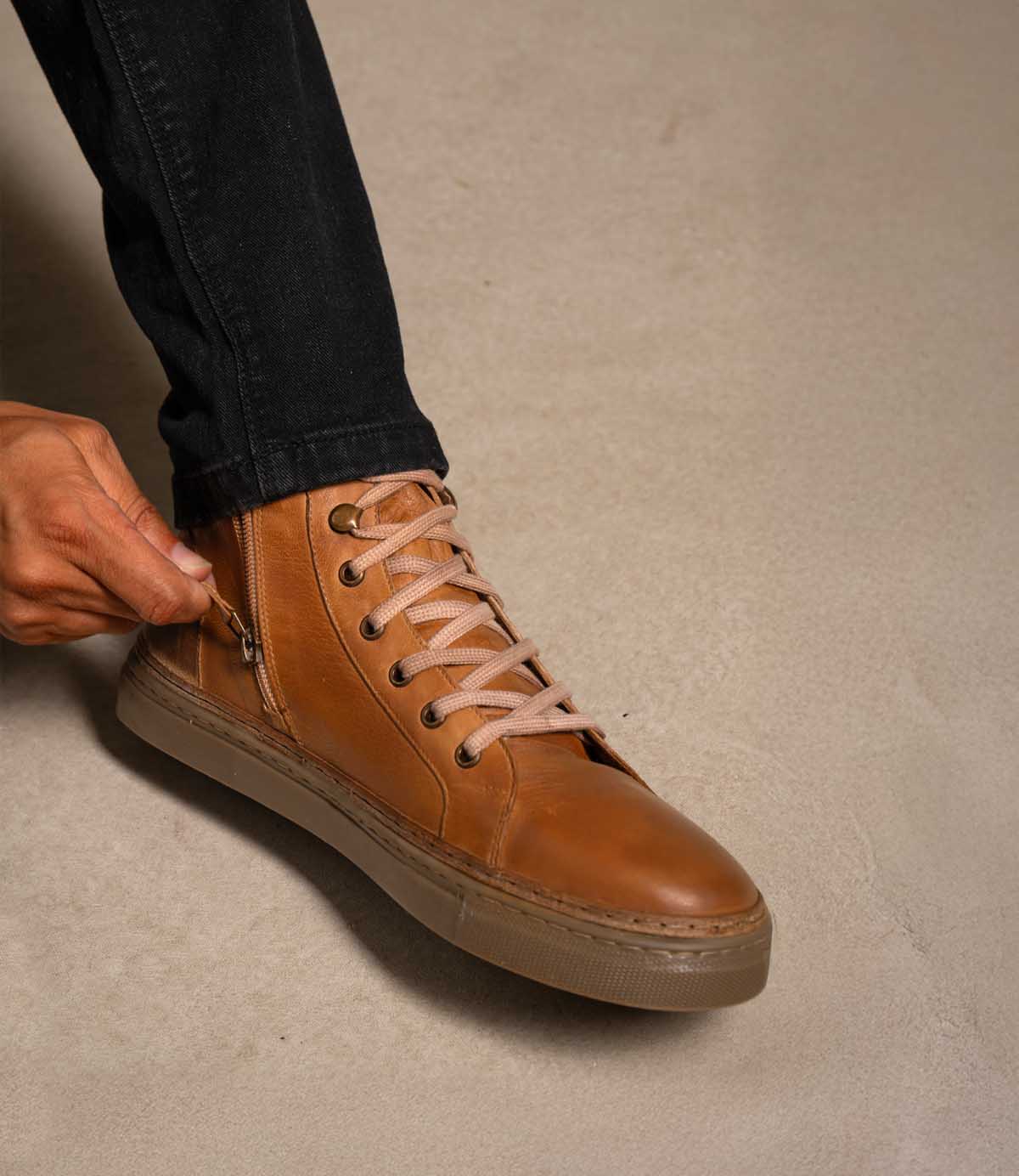 
                  
                    A person wearing black pants zips up a pair of Roan Mac II brown high-top leather sneakers with a padded collar on a beige surface.
                  
                
