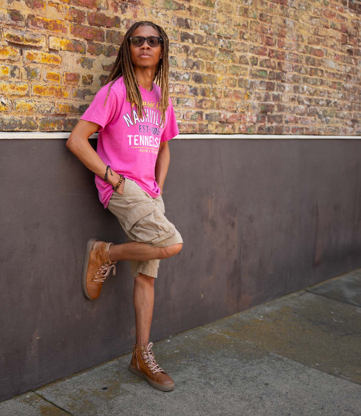 
                  
                    A person with long hair in sunglasses, a pink Nashville tee, khaki shorts, and Roan Mac II high-top leather sneakers with padded collar leans against a brick wall outdoors.
                  
                