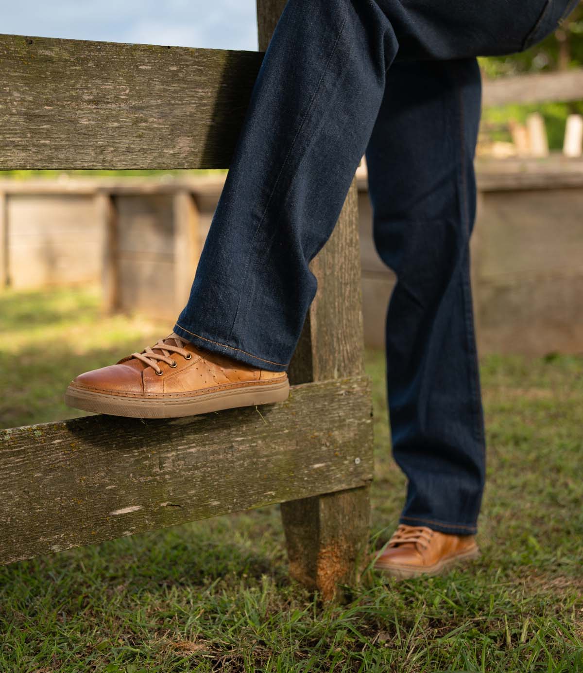 A person wearing blue jeans and Roan Mac II tan high-top leather sneakers with a padded collar rests one foot on a wooden fence outdoors on grass.