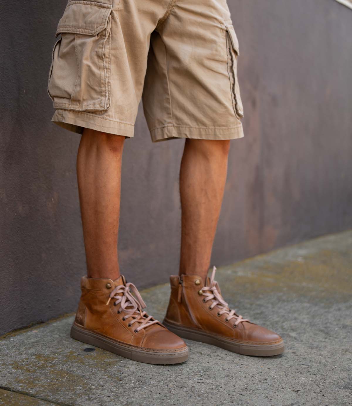 A person stands on a concrete sidewalk next to a gray wall, wearing Roan's Mac II tan cargo shorts and brown high-top leather sneakers with padded collars.