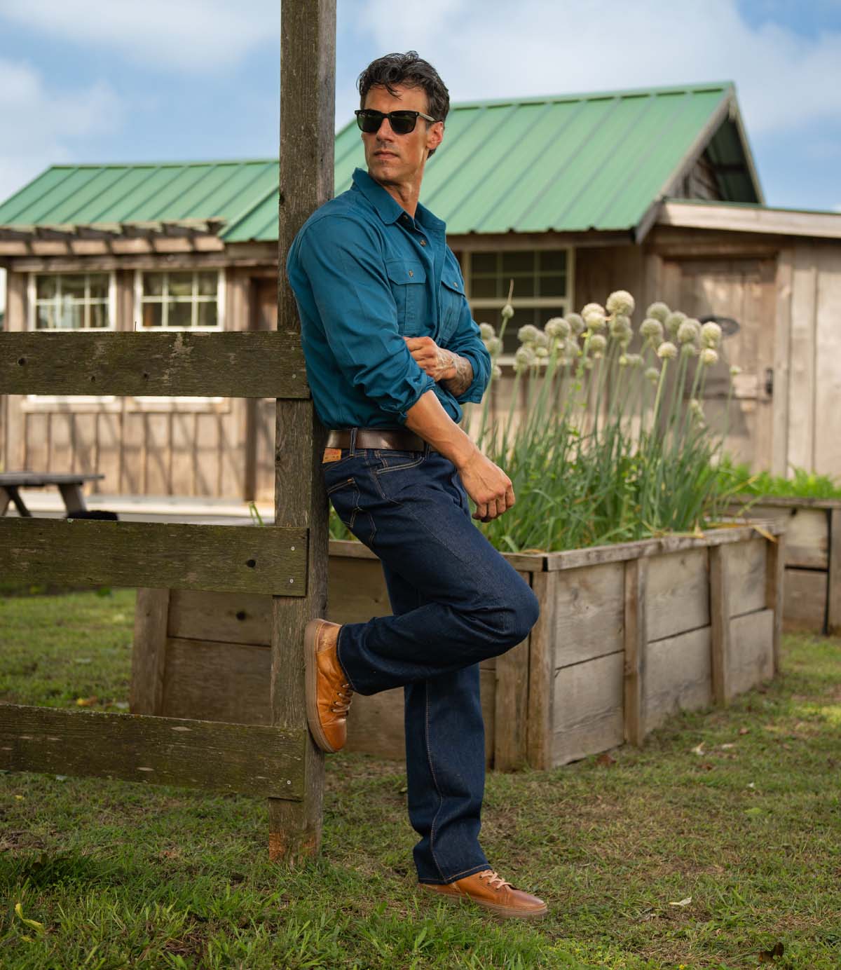 
                  
                    A man wearing Roan's Mac II sunglasses, a teal shirt, blue jeans, and high-top leather sneakers with a rubber outsole leans against a wooden post outdoors near a wooden building and raised garden beds.
                  
                