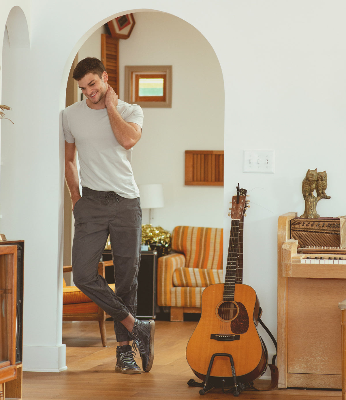 
                  
                    A man in a white t-shirt and gray pants, wearing high-top sneakers, stands smiling under an archway. Nearby are an acoustic guitar, piano, retro armchair, and wall decor. Brand Name: Roan; Product Name: Mac II.
                  
                