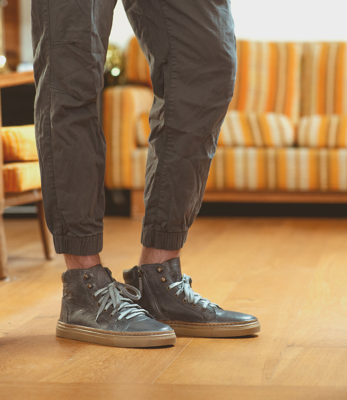 A person wearing Roan's Mac II gray high-top sneakers and dark pants stands on a wooden floor. A striped sofa in the background subtly hints at their outdoor life flair.