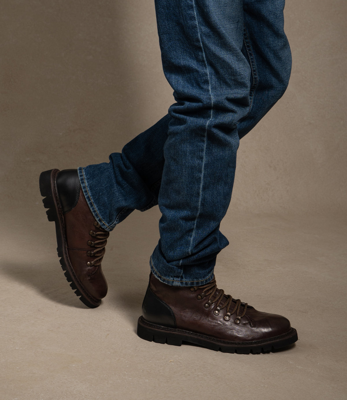 
                  
                    A person wearing dark blue jeans and Roan’s Main brown leather lace-up boots with hiker-style laces, crafted from Alicante leather, stands on a beige floor.
                  
                
