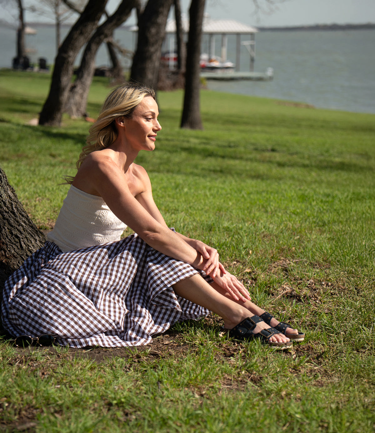 A woman in a white top and checkered skirt sits on the grass by a lake, wearing Roan's Maze suede slip-on sandals and vintage-style studs on a sunny day.