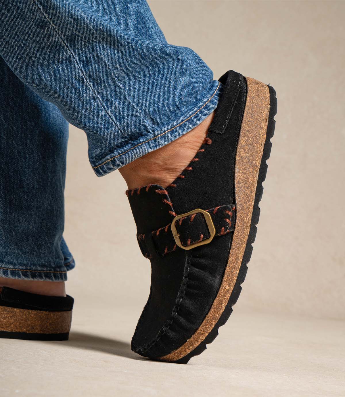 
                  
                    A person wearing blue jeans and the Roan Mingle black plush suede mule slip-on shoes, featuring a chunky cork sole, black outsole, decorative stitching, and a gold-tone buckle detail.
                  
                