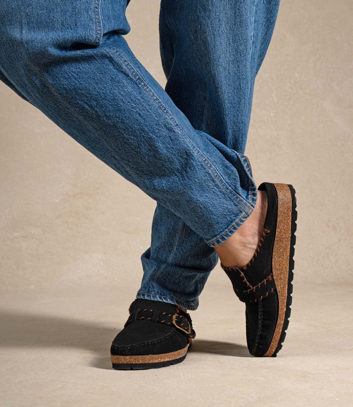 
                  
                    A person wearing blue jeans and the Roan Mingle plush suede mule, detailed with a gold-tone buckle, brown stitching, and a chunky cork sole, stands with legs crossed on a neutral background.
                  
                