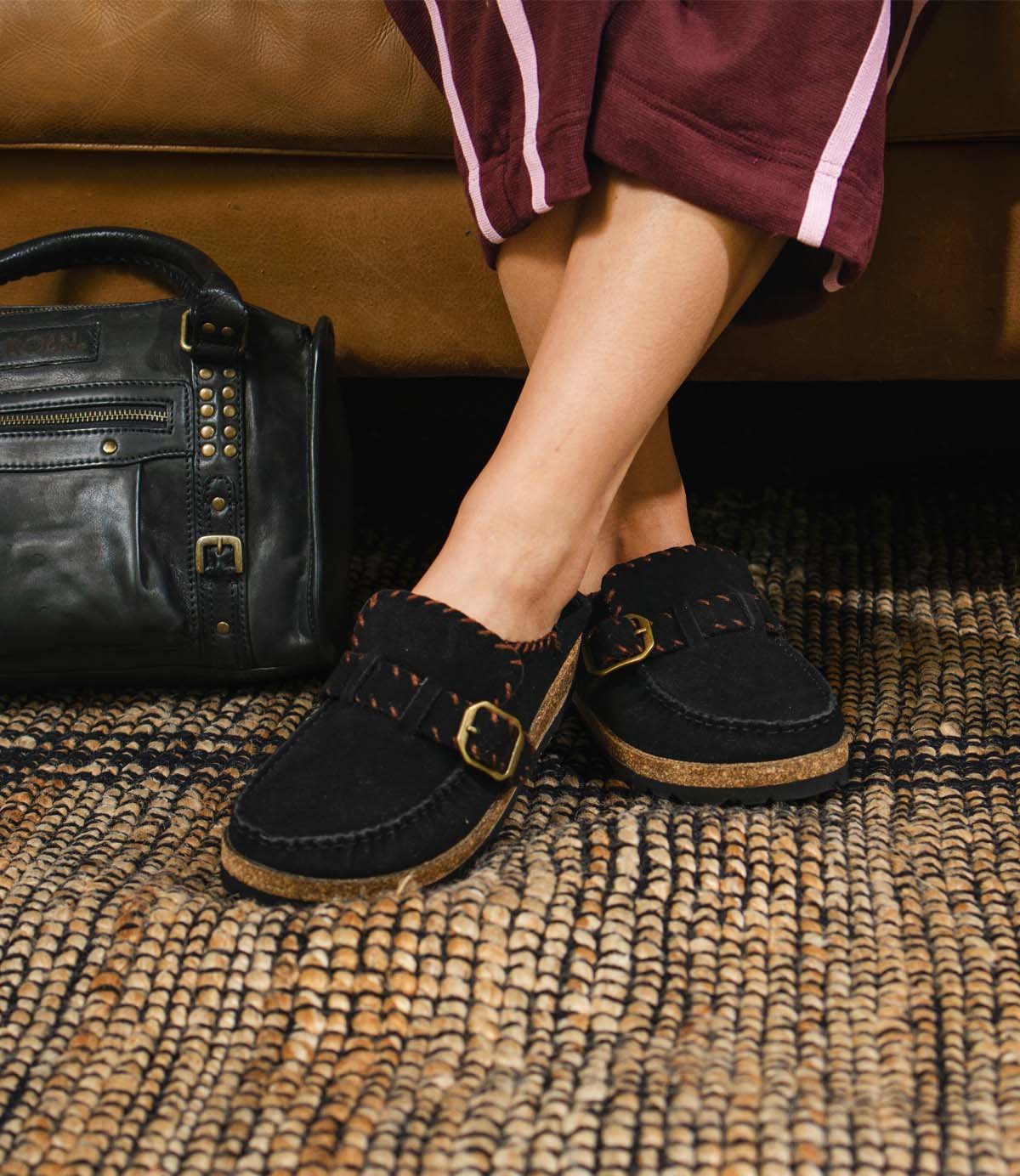 
                  
                    A person wearing black suede Roan Mingle mules with gold-tone buckles and contrast stitching, with a matching black handbag nearby.
                  
                