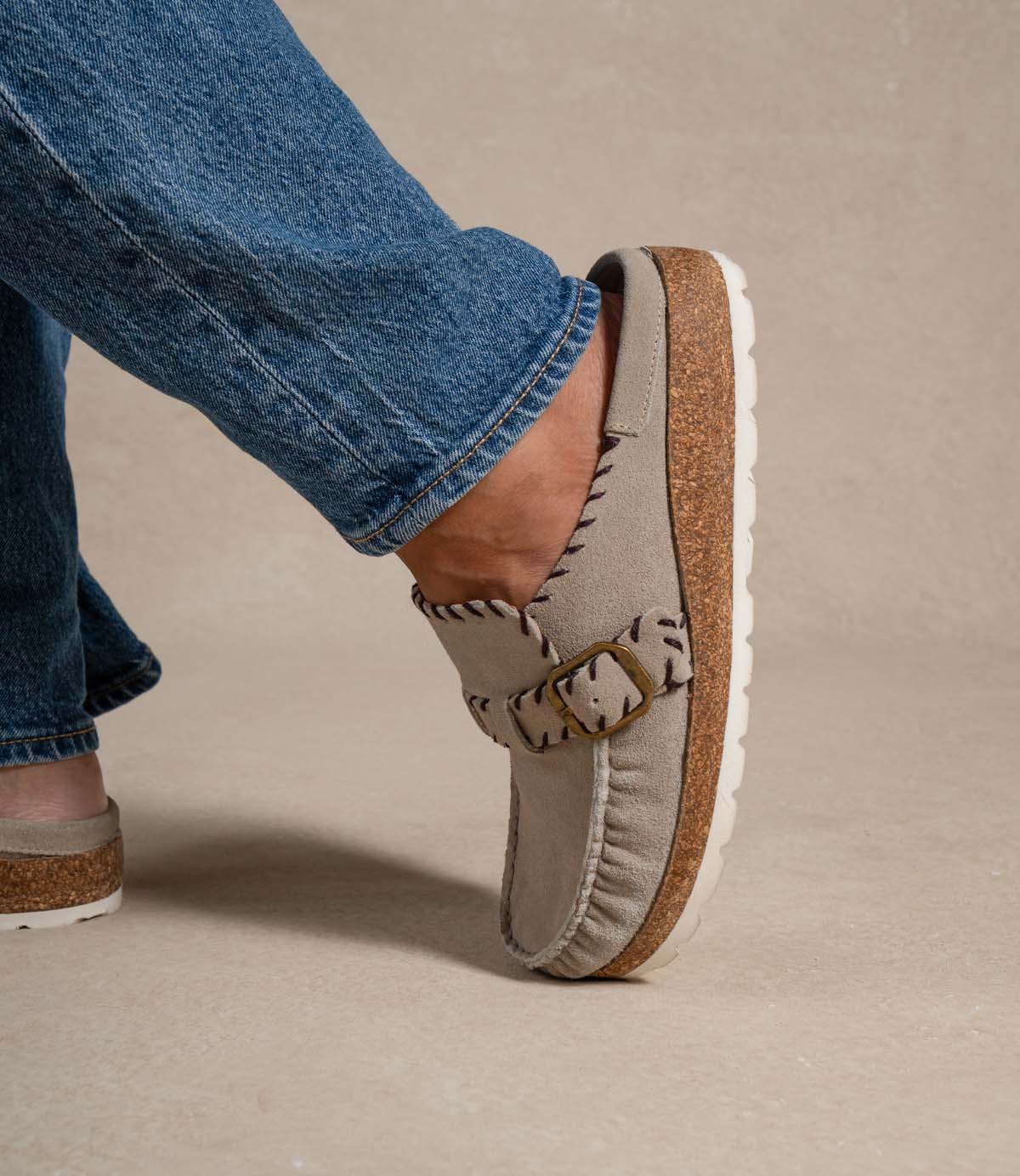 A person wearing the Roan Mingle plush suede mule, featuring a gold-tone buckle and chunky cork sole, paired with blue jeans and standing on a beige surface.