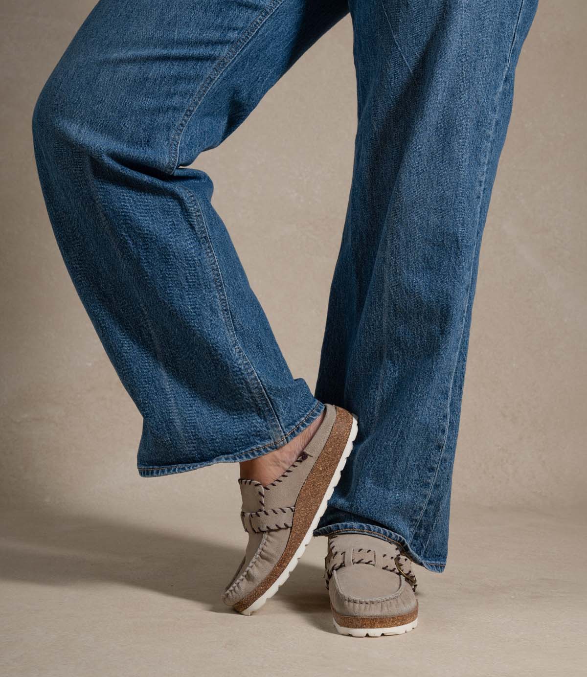 
                  
                    A person wearing blue jeans and the Roan Mingle plush suede mule—featuring a chunky cork sole and gold-tone buckle—stands with one foot raised in a neutral indoor setting.
                  
                
